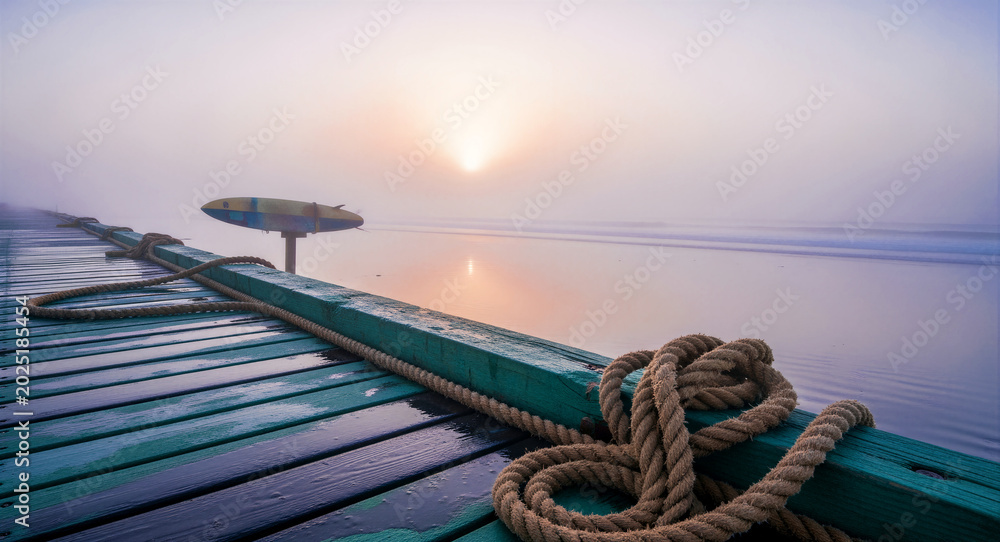 Obraz premium Nautical ropes resting on a wooden pier overlooking a misty beach at sunrise. Soft morning light illuminates the fog over calm ocean waves. Coastal serenity concept, ideal for travel marketing.