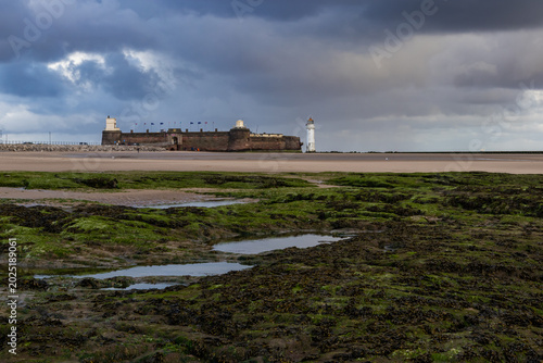 Photos Fort Perch Rock and Perch Rock Lighthouse seen across a seaweed-covered rocky fo