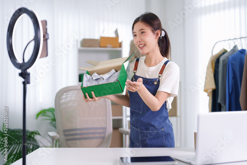 young Asian women entrepreneur live streaming online selling product unbox parcel package ecommerce business from home office studio with ring light smartphone