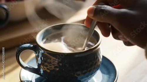 Close-up of a hand stirring hot coffee in a ceramic mug with a spoon.
