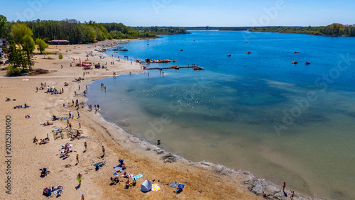 Konstfotografi Aerial drone view of a sandy beach by Niedziegiel lake in Skorzecin, featuring sunbathing people, clear turquoise water, a wooden pier, pedal boats, surrounding green forest, and a clear blue sky
