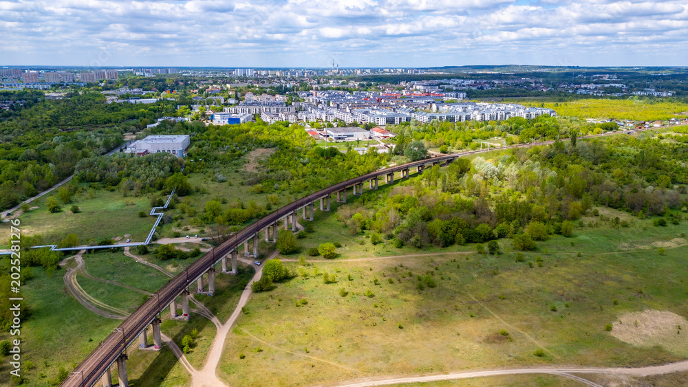 Fototapeta premium Aerial drone view of a long concrete railway viaduct in Poznan stretching across green fields and trees, with a modern city residential district and urban landscape in the background under a cloudy sk