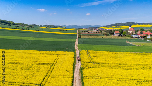 Wide aerial drone view of a farm tractor driving on a dirt road that divides vibrant yellow blooming rapeseed fields and green crops, with a village, church, and hills in the distance.