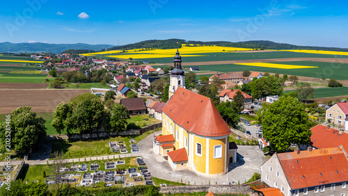 Tableau sur toile Scenic aerial drone view of a historic yellow church with a red tiled roof and adjacent cemetery, situated in a rural village landscape surrounded by blooming rapeseed fields under a blue sky