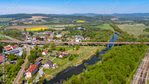 Wide aerial drone view of a long road viaduct and railway tracks crossing over the Nysa Klodzka river near Klodzko, featuring a rural village panorama, green fields, forests, and hills on the horizon.
