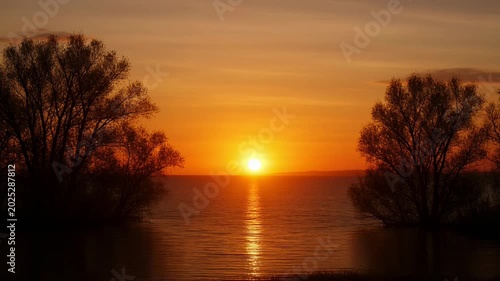 The video captures a serene sunset over a body of water, with the scene gradually transitioning from a bright, glowing sun to a more subdued, reddish horizon