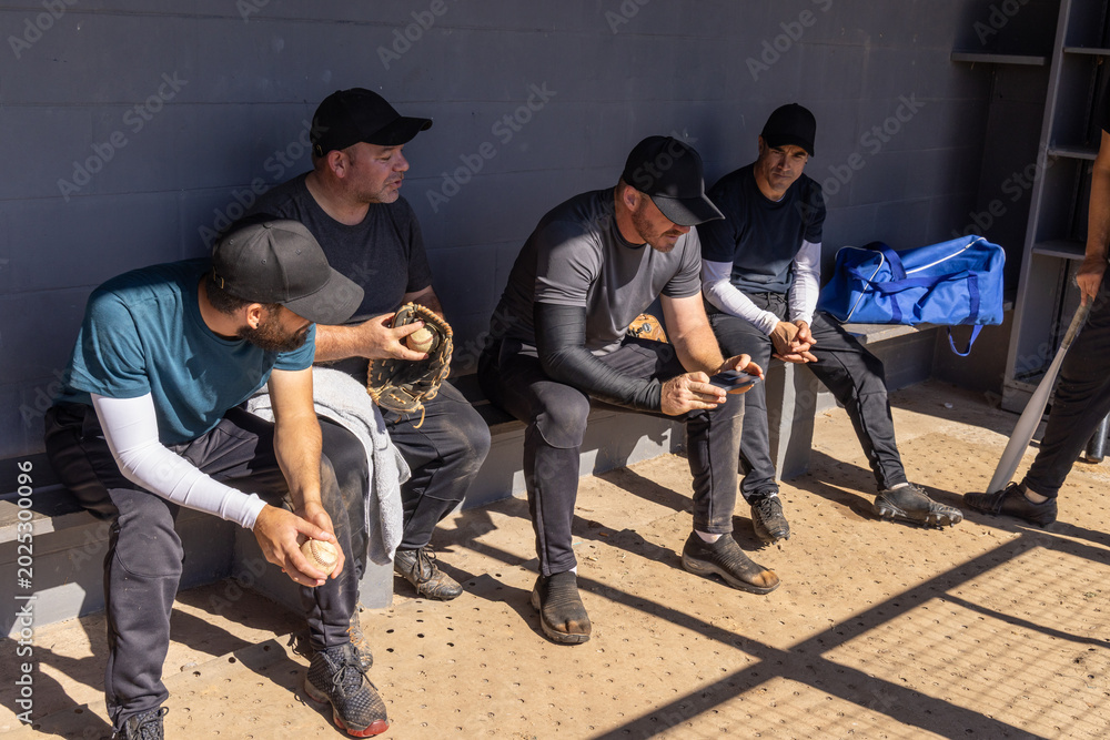 Fototapeta premium Five male teammates wearing caps, sitting on bench in dugout, holding baseballs, using smartphone
