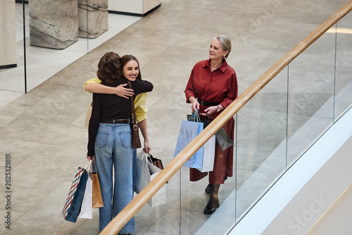 Obraz na plátne Wide high angle shot of Caucasian young women hugging after shopping while senior woman carrying bags approaching in mall, showing family bonding and retail leisure
