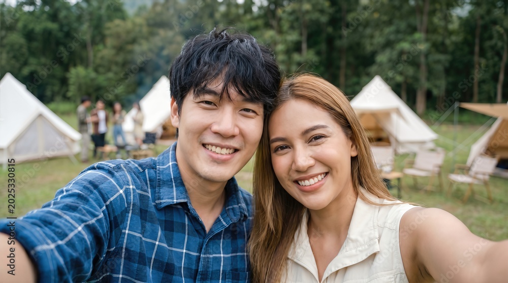 Naklejka premium Young couple taking a selfie in front of camping tents in a forested area, surrounded by friends enjoying outdoor activities and nature