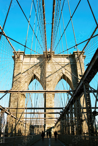 Brooklyn bridge view from pedestrian walkway under stone arches with diverse adults silhouetted at golden hour against clear blue sky urban architectural landmark atmosphere