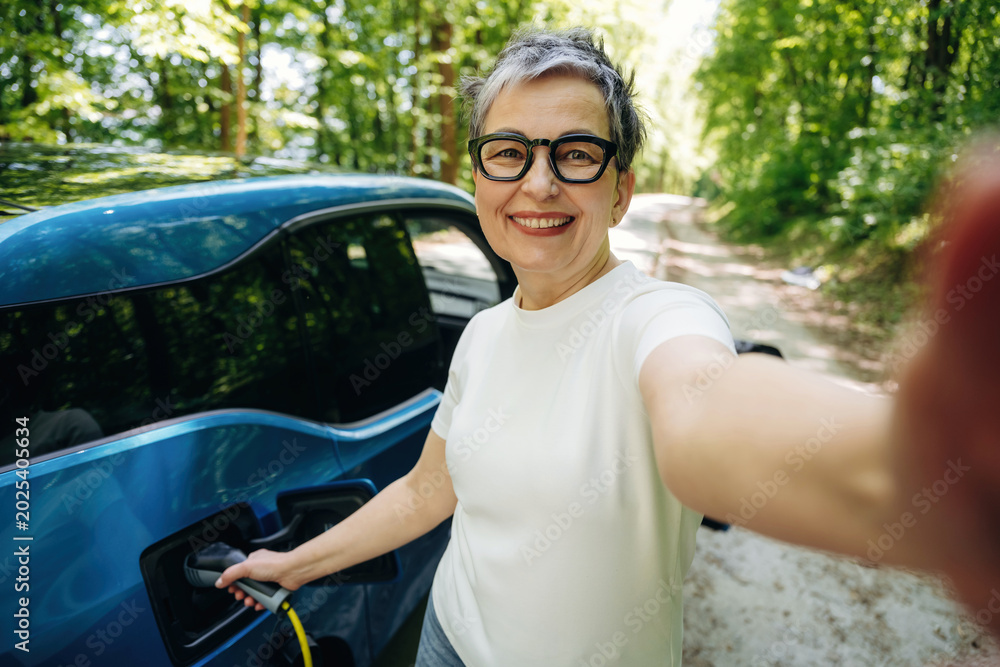Naklejka premium Senior woman with short gray hair and glasses smiles while charging an electric vehicle on a forest road, surrounded by lush green trees and sunlight filtering through