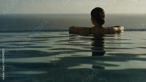 Gazing woman wearing dark straps relaxing on infinity pool edge sea, sun and breeze moving ripples