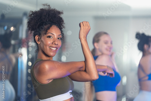 Confident young Black woman with curly hair smiles brightly while flexing her biceps muscle in an olive sports bra, with friends reflected softly in the gym.