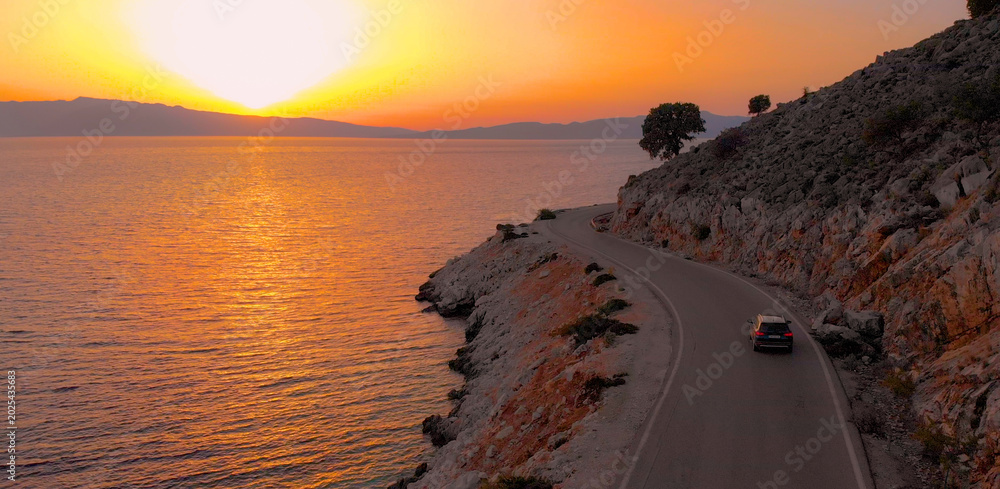 Obraz premium DRONE: Flying behind a big gray car driving along the coastal road at sunrise.