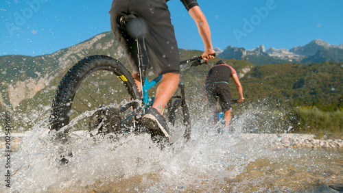 Cuadro en lienzo LOW ANGLE: Glassy drops of water splash at camera as guys cycle across a stream