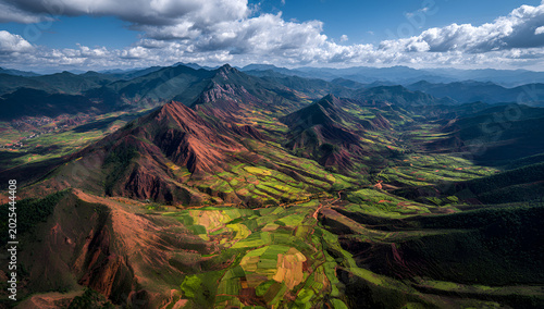 Aerial view captures a dramatic landscape. Rugged red-brown mountains give way to verdant valleys under a bright, cloud-filled sky