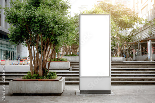 An outdoor advertisement panel stands tall beside trimmed trees in a modern urban landscape. Stairs lead the eye