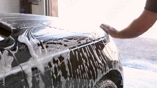 Car wash, close up of male hand wiping black car with sponge full of foam at indoor station. Soap texture and smooth strokes remove dirt, professional auto detailing and cleaning service