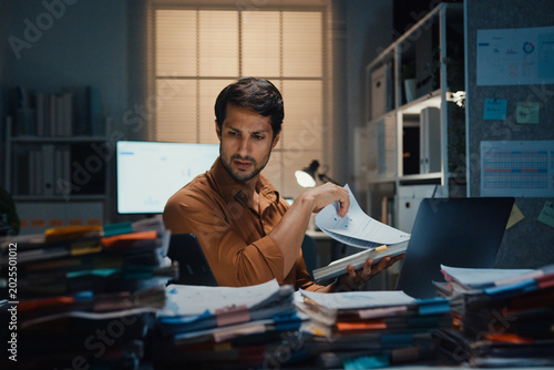 Overworked young Latin businessman working late on document in large stack of paperwork files at office night. Tax audit deadline, accounting stress, corporate burnout, urgent business concept.