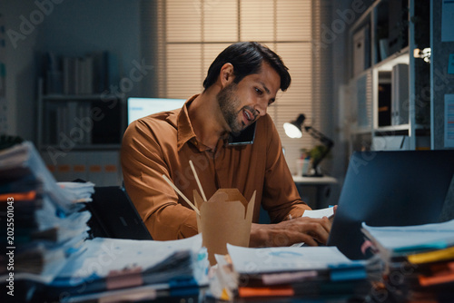 Young Latin businessman taking urgent phone call while reading accounting document on laptop at desk in dark night office. Multitasking overtime, overworked deadline, tired paperwork rush concept.