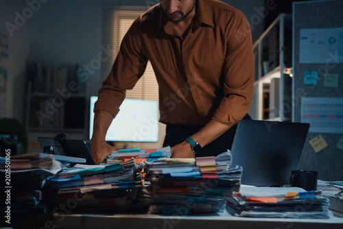Stressed young Latin businessman walking to place tax document stack at desk in dark night office. Overworked overtime, heavy workload stress, tired employee, corporate business burnout concept.