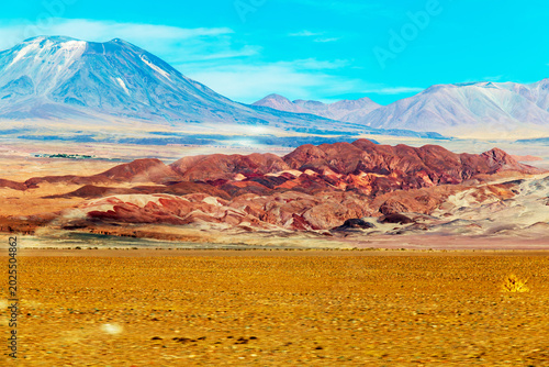 Colorful desert hills rise toward snow‑capped Andean peaks under a bright sky in the Atacama Desert of northern Chile