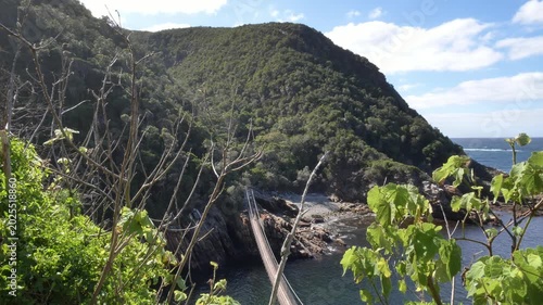 A breathtaking aerial view of a partial suspension bridge, rugged cliffs, the ocean, and lush forests creates a dramatic landscape at Storm’s River Mouth in Tsitsikamma National Park, South Africa.