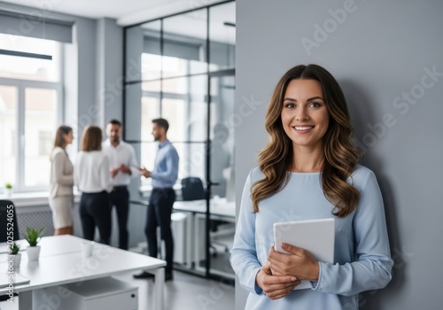 Professional woman holding a tablet in a modern office environment with colleagues in the background
