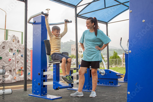 Female instructor helping young child during a fitness workout in outdoors gym. Children fitness training and  healthy lifestyle concept