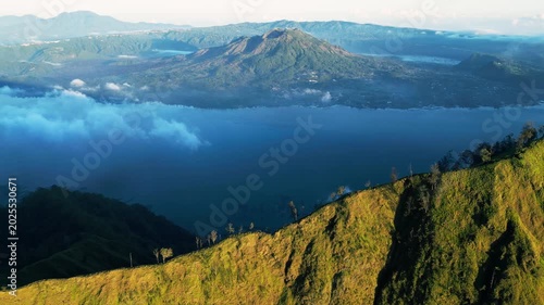 Volcanic caldera lake in Indonesia with crater landscape and steep mountain slopes, sunlit terrain with clouds and Mount Batur in the background, push-in aerial drone footage