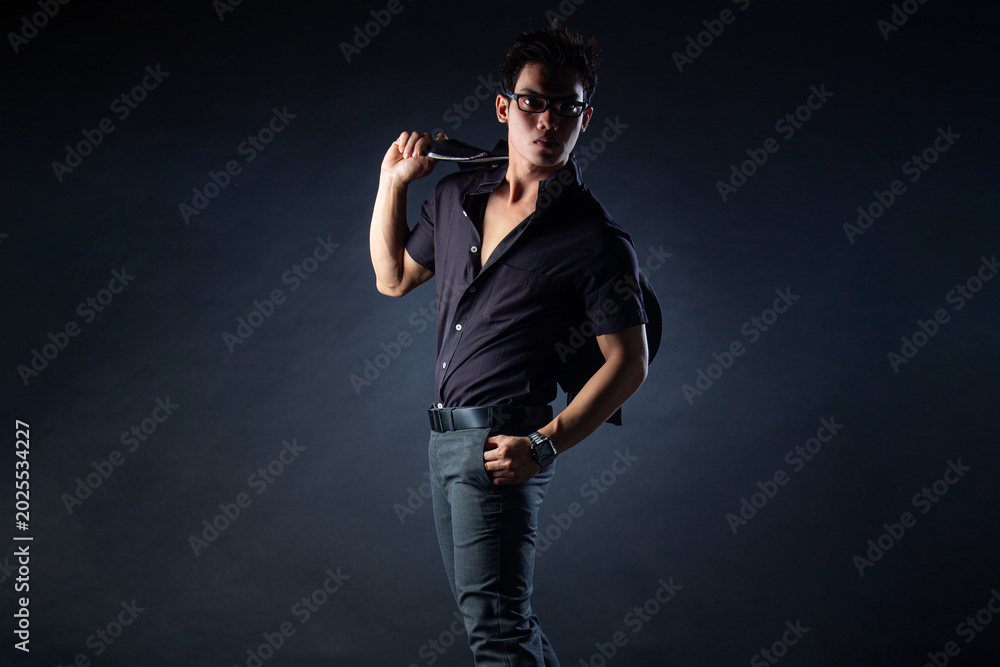 Fototapeta premium An Asian man in all-black formal attire poses against a dark background with dramatic studio lighting, holding his jacket and wearing stylish glasses.