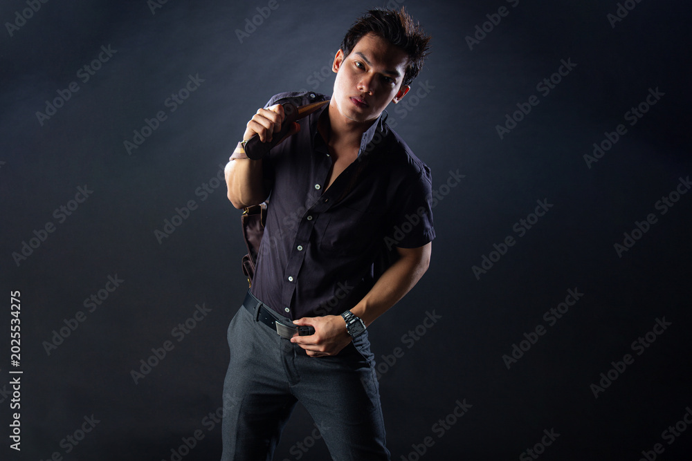 Fototapeta premium An attractive Asian man dressed in all-black formal attire poses confidently in a dramatic studio setting, holding a stylish brown duffle bag with a strong and dynamic stance against a dark background