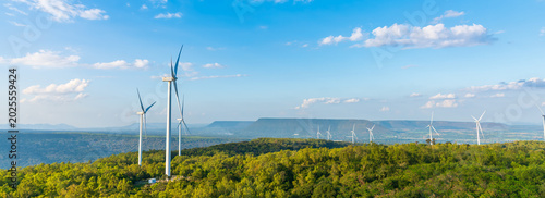 Panoramic view of a large wind farm with turbines on green hills. Renewable energy production and clean technology landscape under blue sky with clouds.