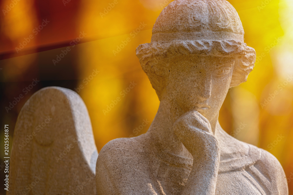 Fototapeta premium Thoughtful angel statue with hand near lips in warm golden sunlight, symbolic image of silence, contemplation and spiritual mystery