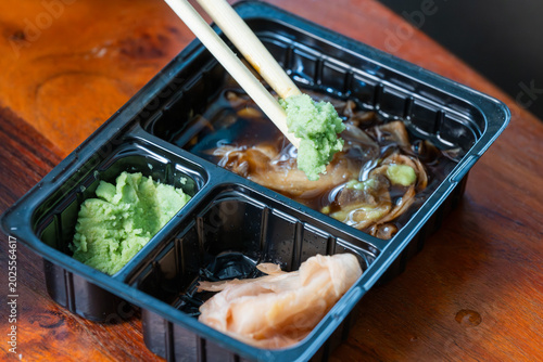 Fotografie wooden chopsticks lifting a dollop of green wasabi from a black plastic takeout container filled with soy sauce and pickled ginger
