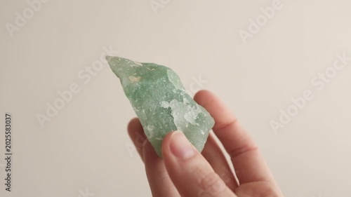 Close-up of a person's hand holding and turning a raw, unpolished green aventurine quartz crystal gemstone against a neutral studio background, horizontal orientation.