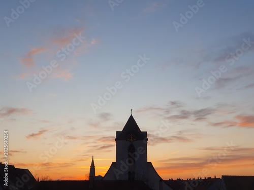 Принти на полотні A peaceful church silhouette and clock tower rise above a quiet town roofline against a colorful sunrise sunset sky, creating a tranquil, contemplative mood with soft clouds and warm evening light