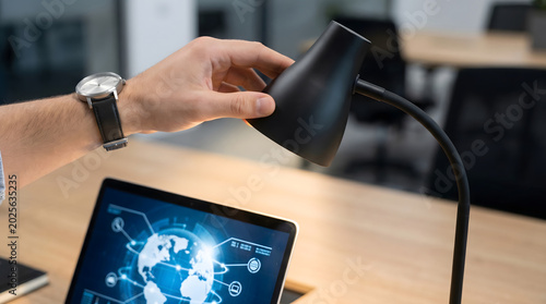 Close Up of Hand Adjusting Desk Lamp Over Laptop Screen Displaying Global Network Tech Interface in Modern Corporate Office Workspace