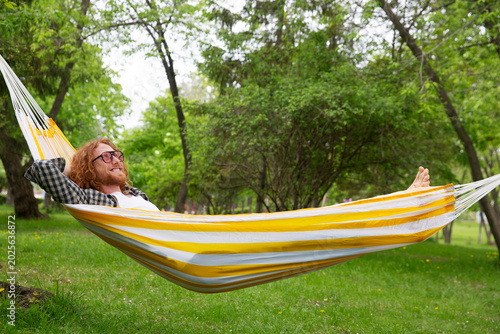 portrait of mature smiling man with red hair in pink glasses lying in a hammock in the garden on a summer day