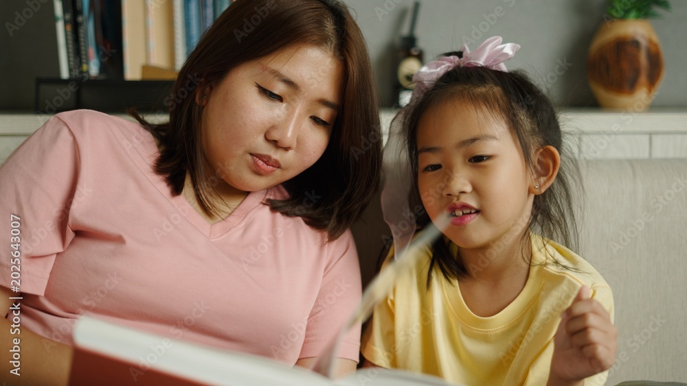 Obraz premium Asian mother and daughter reading book together at home, learning and bonding