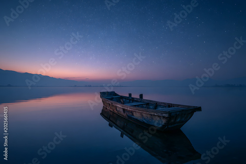 Old wooden boat on still water at twilight, with a starry sky, colorful horizon, and reflections creating a serene and tranquil atmosphere.