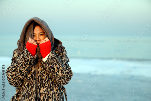 Εκτύπωση καμβά Winter seaside portrait: lady on sandy beach under hood, red cuffs and green nails accentuating enigmatic oceanfront expression