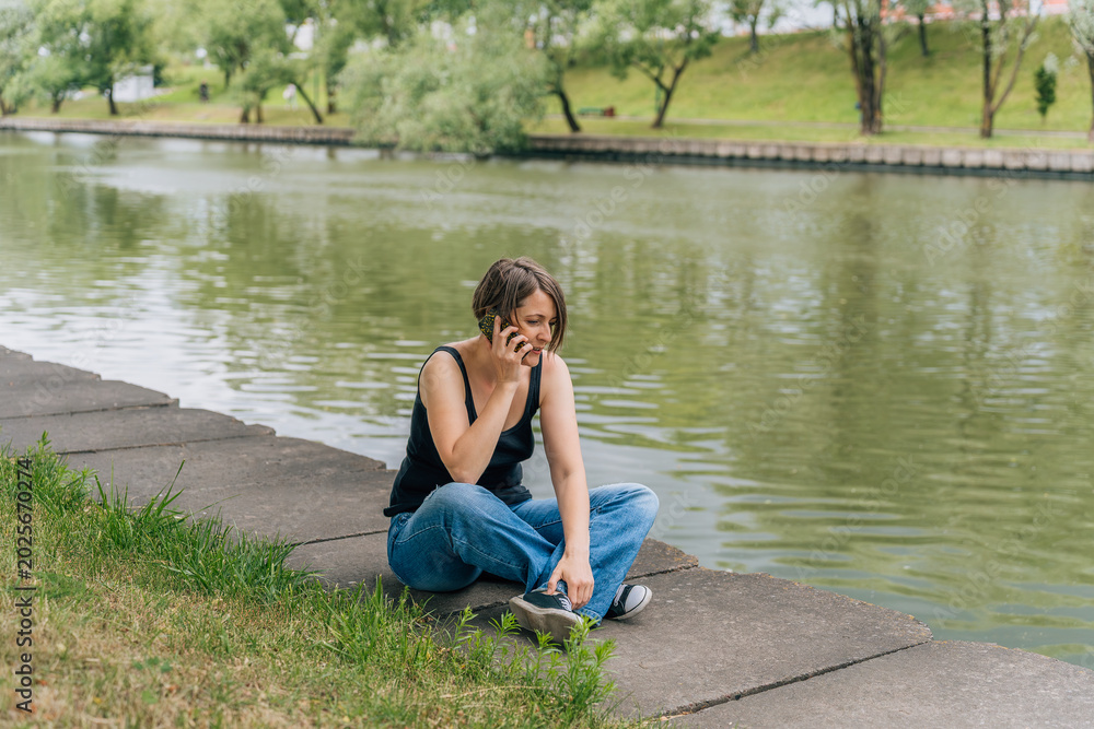 Fototapeta premium Woman 40s talking on phone while sitting on urban riverbank