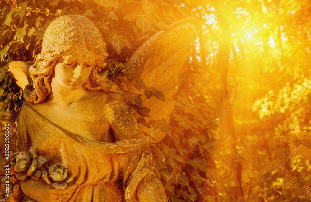 Fototapeta premium Golden angel statue glowing in warm sunset light among forest trees