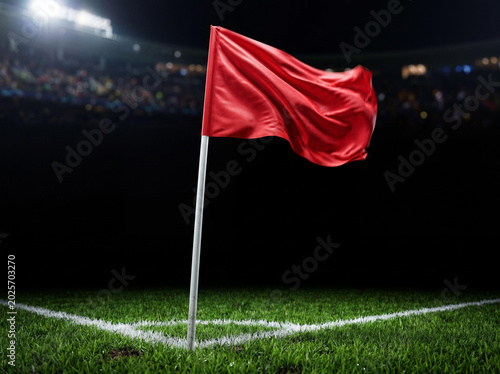 Red corner flag waving on a vibrant green soccer field corner at night, brightly lit by stadium lights with a blurred crowd in the distant background, on black background.