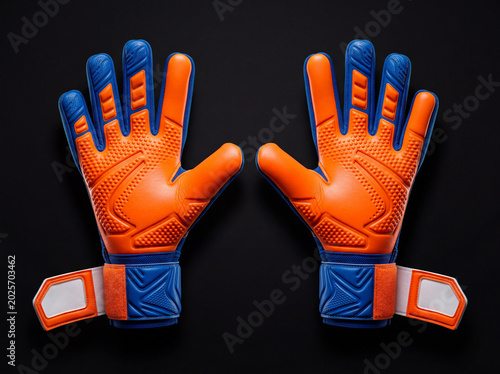 Pair of new soccer goalkeeper gloves, vibrant orange and blue with textured palms and open wrist straps, sporting equipment on black background.
