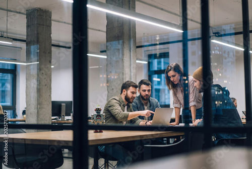 Creative startup team collaborating on a laptop in a modern glass office with a robot toy on the desk, viewed through a partition