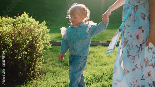 Young child walks beside an adult, holding hands. They move slowly along a garden path. Sunlight glows warmly around them. Green grass and bushes surround the scene. The child looks focused