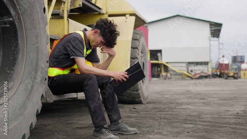 Foto A stressed engineer reviewing a difficult report at an industrial site, sitting on heavy machinery and showing signs of workplace burnout and pressure
