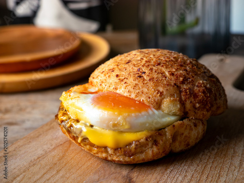 Rustic sourdough bread roll sandwich with sunny side up fried egg and melted cheese on wooden cutting board, homemade breakfast brunch